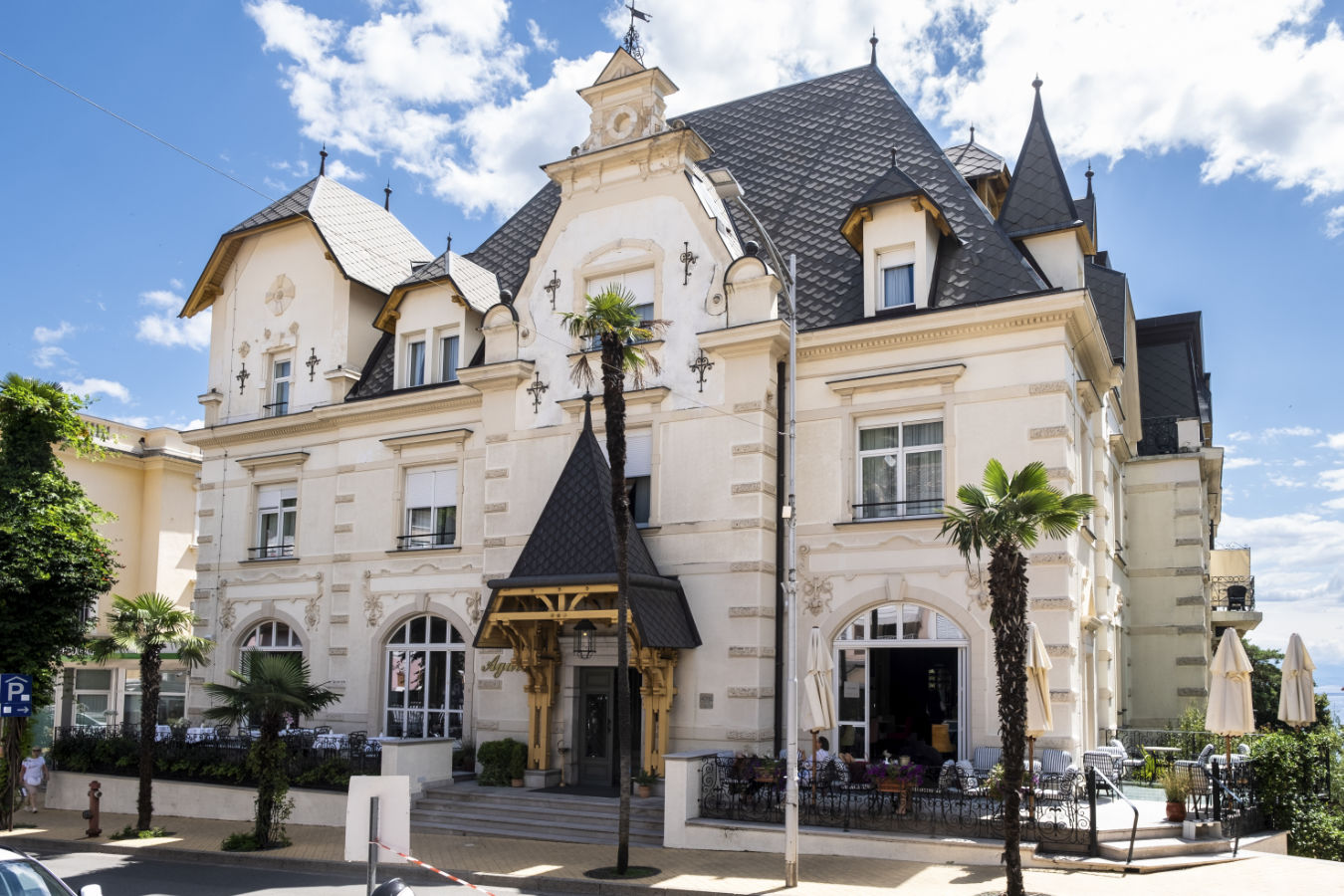 Grand bâtiment blanc de plusieurs étages avec un toit en ardoise et des détails architecturaux, l'hôtel Agava à Opatija dispose de charmants sièges en plein air au rez-de-chaussée, sous un ciel bleu limpide. Des palmiers encadrent la scène accueillante de cet hôtel pittoresque en Croatie.