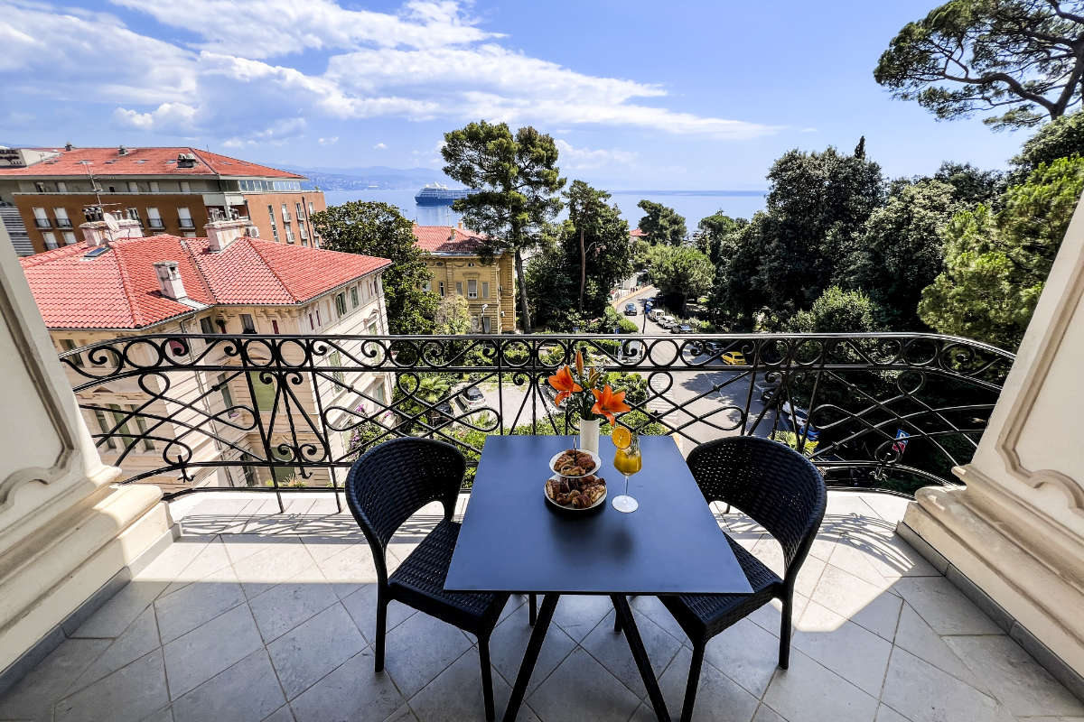 Una vista dal balcone dell'Hotel Agava Opatija mostra un piccolo tavolo nero con un vaso di fiori, una ciotola di snack e un bicchiere. Due sedie nere circondano il tavolo. Sullo sfondo ci sono edifici, alberi e una vista lontana del mare.