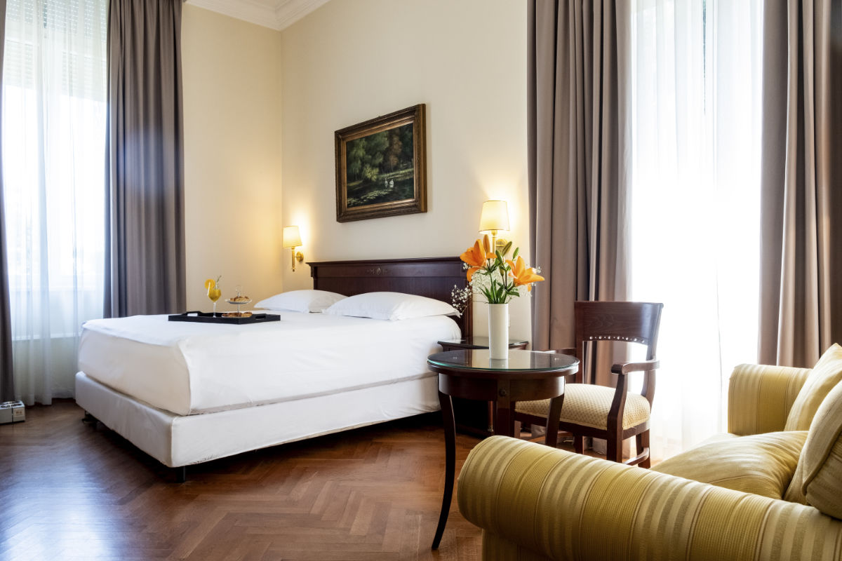 Una camera d'albergo dell'Hotel Agava, a Opatija, con un letto matrimoniale, un tavolo rotondo ornato di fiori, una poltrona e ampie finestre coperte da tende. Sopra il letto è appeso un quadro e vi è appoggiato un vassoio con degli oggetti.