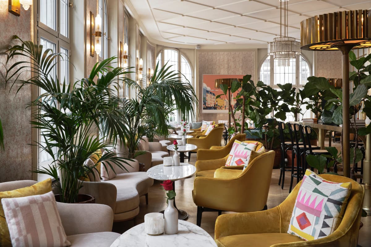 Chic lounge at Hotel Capital with yellow and beige armchairs, patterned pillows, marble tables, potted plants, large windows, and modern lighting fixtures in a bright, stylish interior—an inviting setting that truly reflects the elegance of one of the best hotels Croatia has to offer.