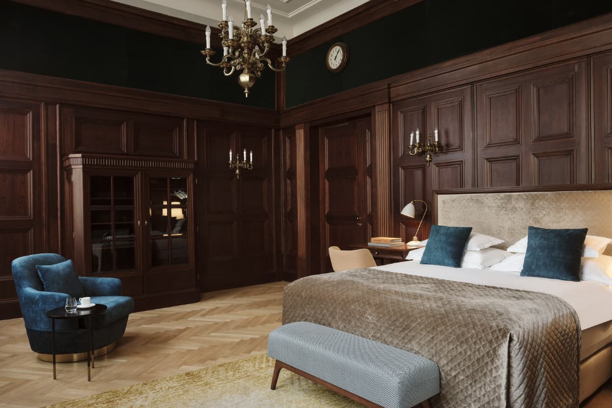 Elegant hotel room at Hotel Capital Zagreb with wood-paneled walls, a large bed with blue pillows, a chandelier, a blue armchair, a clock, and herringbone wood floors—experience one of the best hotels Croatia has to offer.
