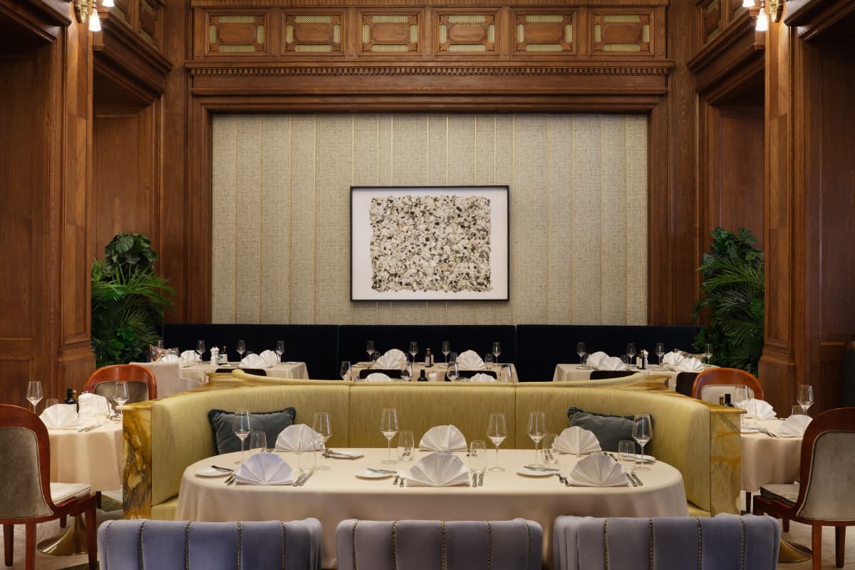 Elegant restaurant dining area with round tables set with white tablecloths and napkins, surrounded by wooden walls, plants, and a framed abstract artwork on the back wall—an inviting setting often found in some of the best hotels Croatia has to offer, such as Hotel Capital Zagreb.