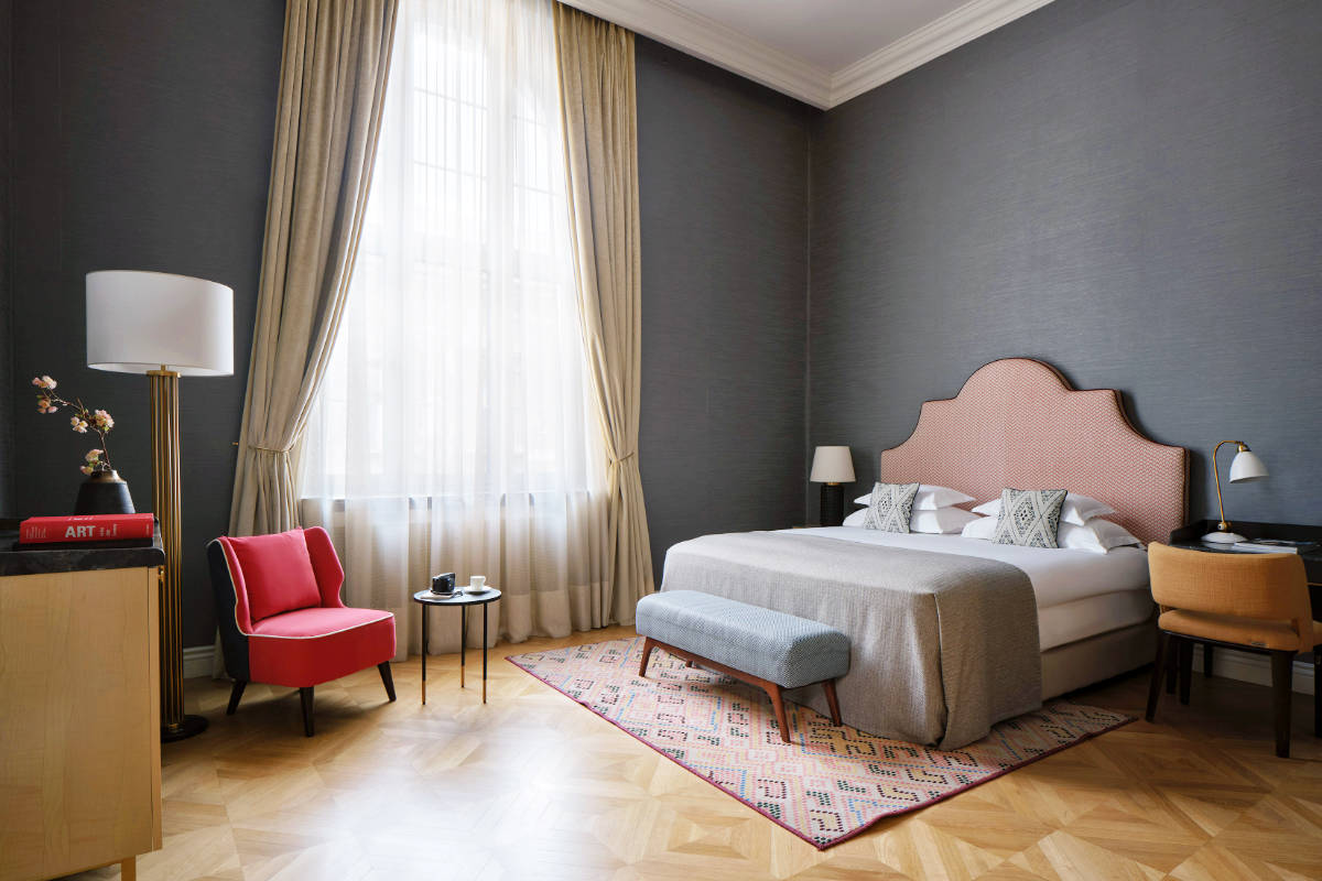 Modern hotel room at Hotel Capital Zagreb, featuring a large bed, bench, red chair, desk, lamp, and a tall window with sheer curtains letting in natural light—an inviting space in one of the best hotels Croatia has to offer.