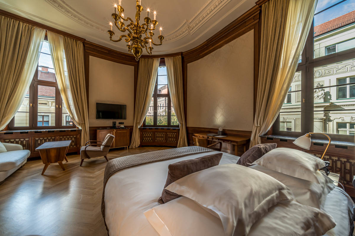 Una lussuosa camera dell'Hotel Capital con un grande letto con più cuscini, alte finestre con tende, mobili in legno, lampadario e vista sugli edifici storici di Zagabria.