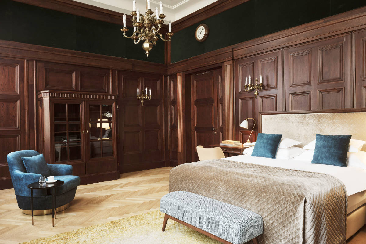 Elegant hotel room with wood-paneled walls, a large bed with blue and beige accents, chandelier, blue armchair, small table, desk, and wall clock—an inviting space that reflects the charm of Hotel Capital Zagreb. Experience timeless comfort in one of the best hotels Croatia has to offer.