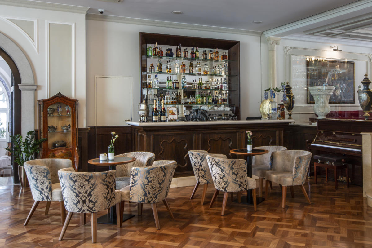 Le bar sophistiqué de l'hôtel Milenij est composé de fauteuils à motifs disposés autour de petites tables, d'une étagère à alcool bien remplie derrière un comptoir en bois et d'un piano en arrière-plan.