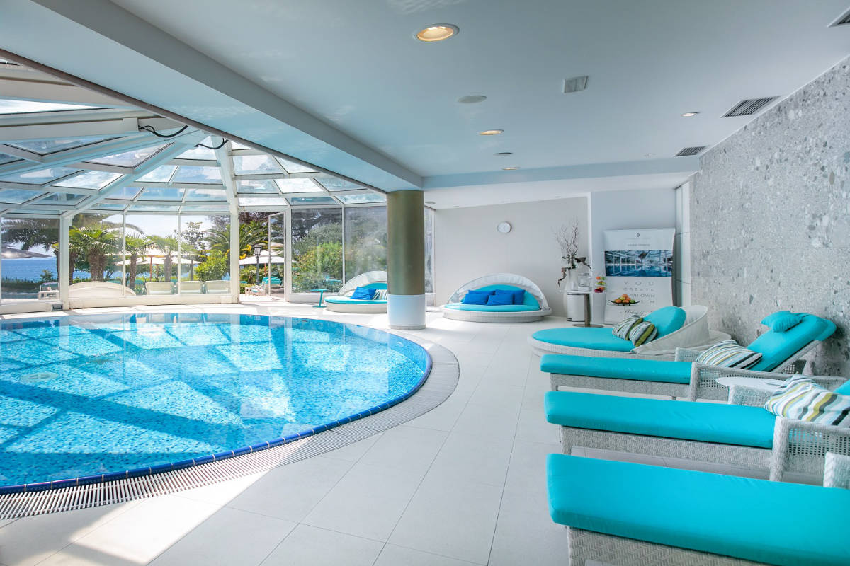 La piscine intérieure de l'hôtel Milenij est équipée de chaises longues turquoise, d'un bassin circulaire et de grandes fenêtres donnant sur une végétation luxuriante. L'espace est lumineux et moderne, avec un décor blanc et turquoise qui crée une atmosphère sereine.