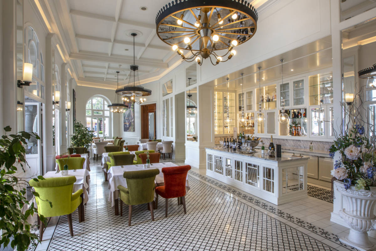 Ce restaurant bien éclairé de l'hôtel Milenij présente des murs blancs, de grandes fenêtres, des chaises aux couleurs vives, un carrelage à motifs et deux grands lustres. Les tables sont recouvertes de nappes blanches impeccables, tandis que le comptoir présente un large éventail de produits.