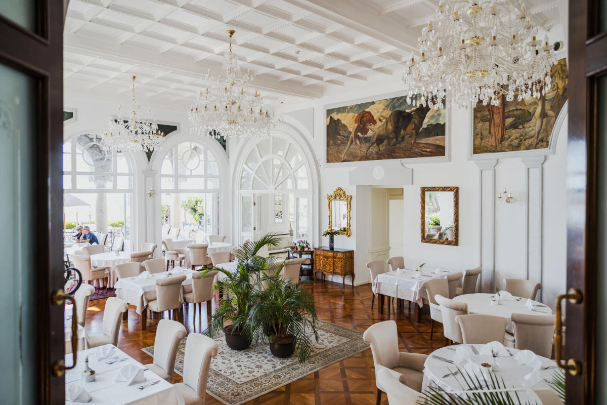 La salle à manger de l'Hotel Milenij, bien éclairée et élégamment décorée, est ornée de lustres, de tableaux et de fenêtres cintrées. Les tables sont recouvertes de nappes blanches et de chaises rembourrées. Des plantes vertes sont disposées près de l'entrée.