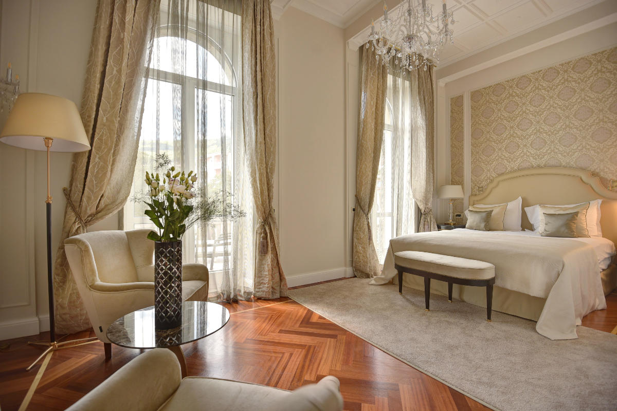 Chambre luxueuse à l'hôtel Milenij avec un grand lit, un décor élégant, un lustre, un fauteuil, une table ronde avec des fleurs, et la lumière naturelle des grandes fenêtres avec des rideaux.