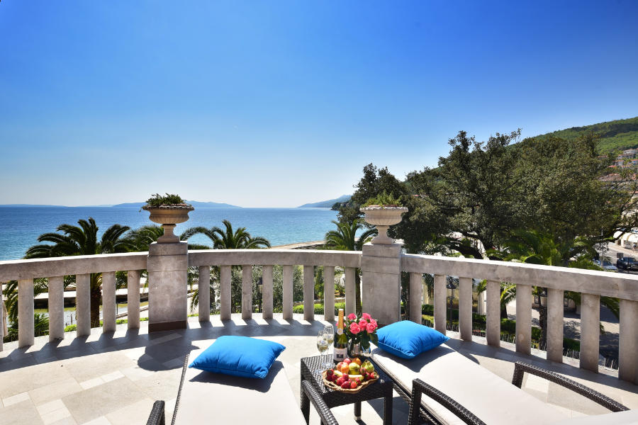 Le balcon de l'hôtel Milenij, situé au bord de la mer, est équipé de deux chaises longues avec des coussins bleus. Une table contient une bouteille de vin, des verres et une coupe de fruits. L'océan et les arbres sont visibles à l'arrière-plan sous un ciel bleu clair.