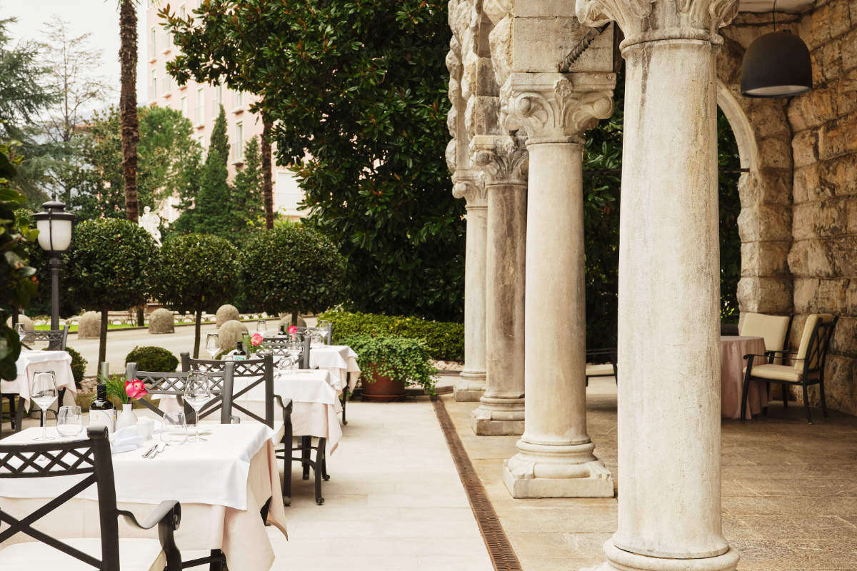 Outdoor restaurant terrace with stone columns, white tablecloth-covered tables, black chairs, and lush greenery in the background—an inviting dining experience characteristic of a 5 star hotel Croatia is renowned for. Enjoy refined cuisine in this elegant setting at one of the best hotels Croatia has to offer, such as Hotel Opatija.