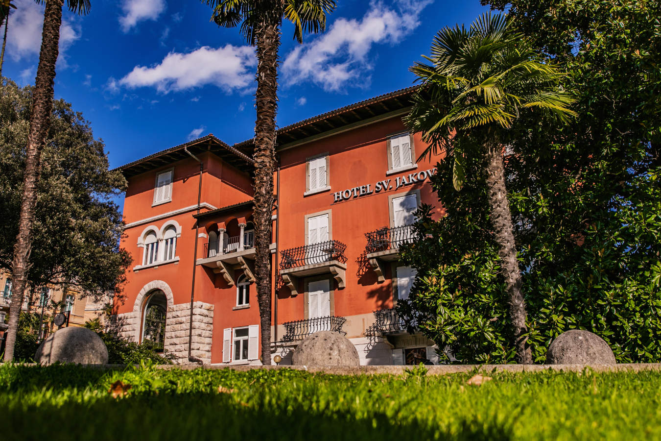 "El Hotel Sveti Jakov está rodeado de exuberante vegetación y palmeras bajo un cielo parcialmente nublado.