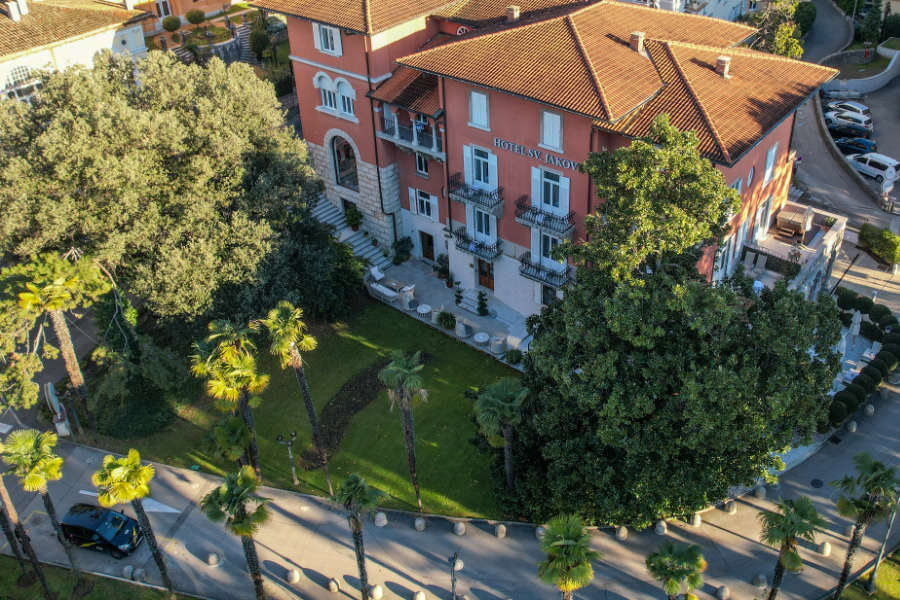 Luftaufnahme des Hotels Villa Naccarati, das von Bäumen und einem gepflegten Garten umgeben ist und an einer von Palmen gesäumten Straße liegt, die an den eleganten Charme des Hotels Sveti Jakov Opatija erinnert.