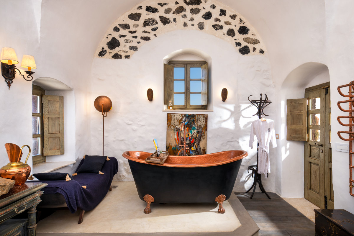 A rustic bathroom in Villa Santa Croce features a black freestanding bathtub, a wooden window, a lounge chair, wall decorations, a freestanding coat rack, and antique accents under an arched, stone-accented ceiling.
