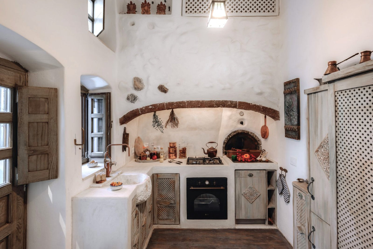 Rustic kitchen in Villa Santa Croce with white walls, an arched oven, wooden cabinets, and various hanging herbs. A window on the left lets in natural light, and the decor includes dried flowers and pottery.