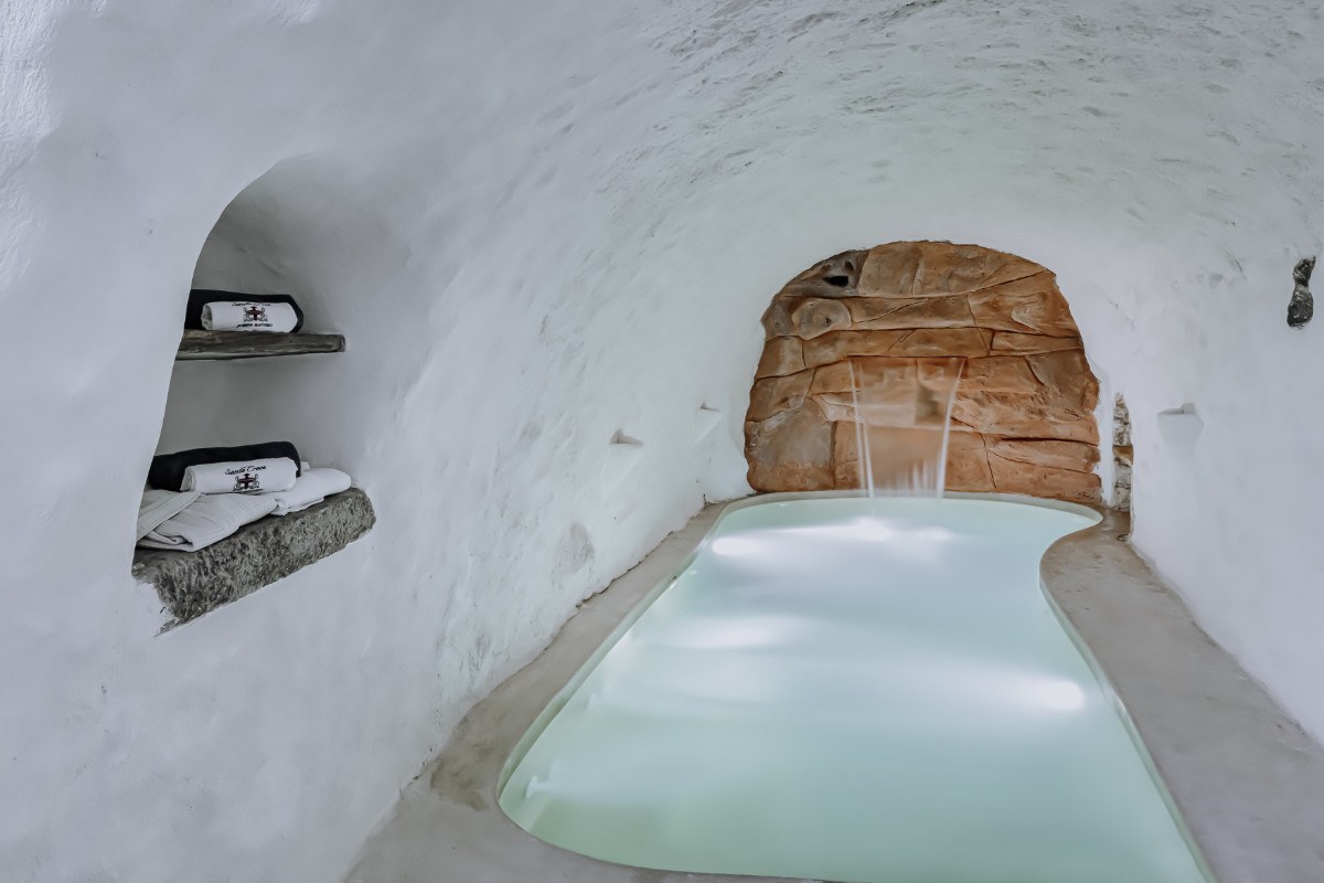 A small indoor pool with white, curved walls and a waterfall feature, reminiscent of the elegance found at Villa Santa Croce. Towels are neatly stacked on shelves carved into the wall.