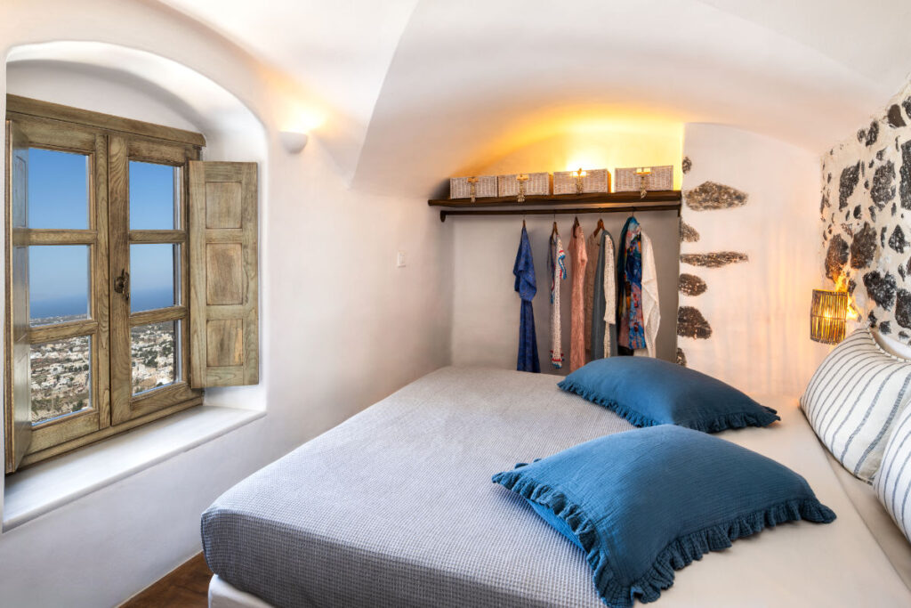 Capolavori in movimento: Le mostre d'arte da non perdere in Europa quest'estate 14 L'accogliente camera da letto di Villa Santa Croce è dotata di un letto con cuscini blu, di una finestra che offre una vista panoramica e di un armadio con diversi abiti appesi illuminati da luci ambientali. Le pareti vantano accenti rustici in legno e pietra, che incarnano il fascino sereno di Villa Santa Croce a Santorini.