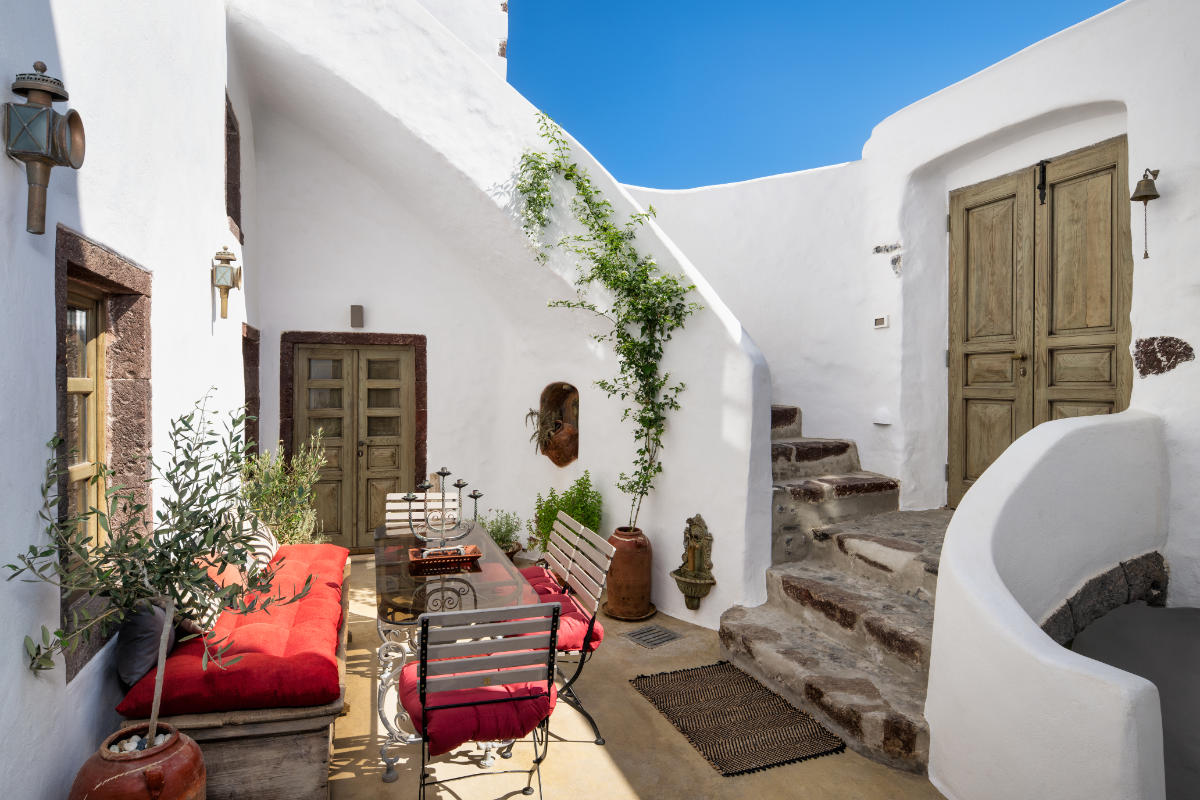 A cozy outdoor courtyard with white walls, a red-cushioned seating area, a dining table, wooden doors, stone steps, and various potted plants evokes the charm of Villa Santa Croce.