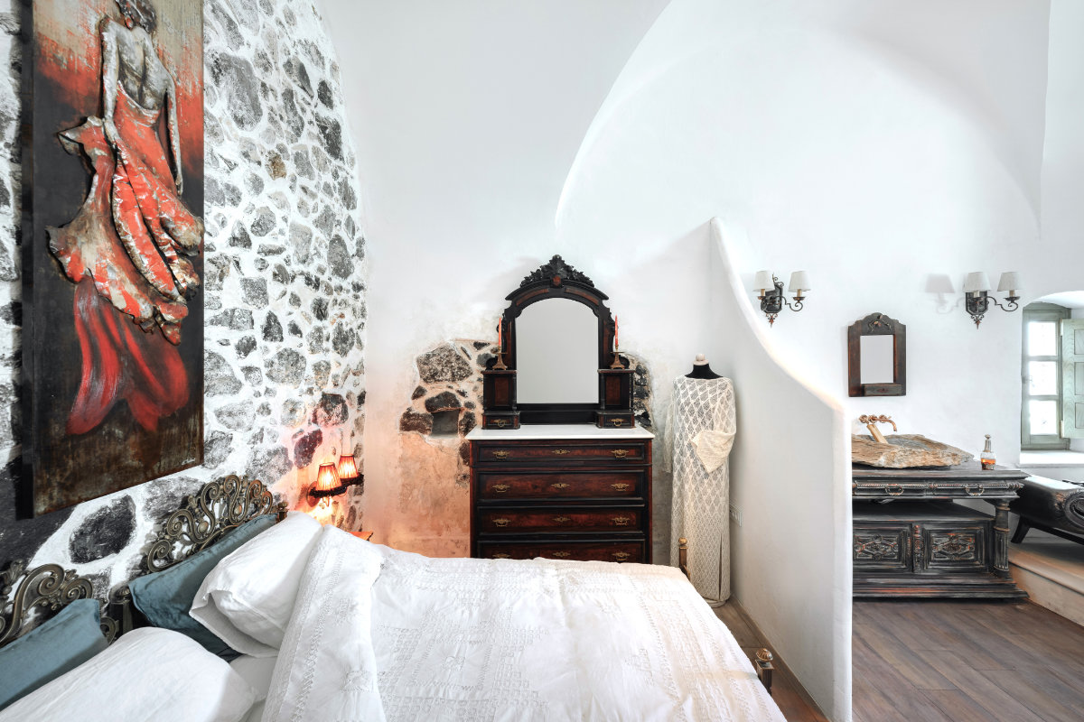 A serene bedroom in Villa Santa Croce features a white bedspread, a stone accent wall, an ornate wooden dresser with a mirror, and a white dress on a mannequin. A painting and various wall accessories complete the elegant ambiance.