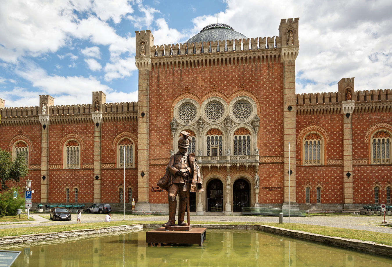 In front of the Museum of Military History, a statue of a figure in armor stands proudly against the historic brick facade with arched windows and decorative elements, its grand form mirrored in the adjacent pond under a partially cloudy sky.