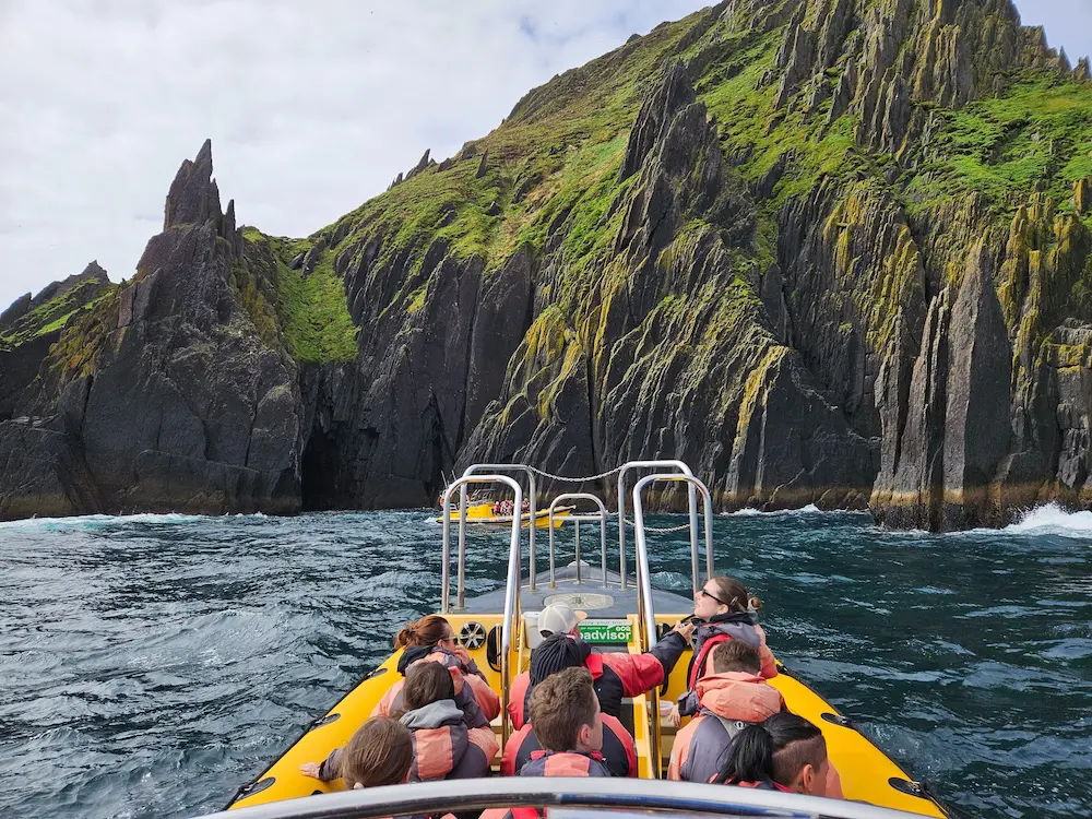 Un grupo de personas con chalecos salvavidas se sientan en una barca amarilla que se acerca a unos acantilados escarpados y rocosos cubiertos de musgo verde.