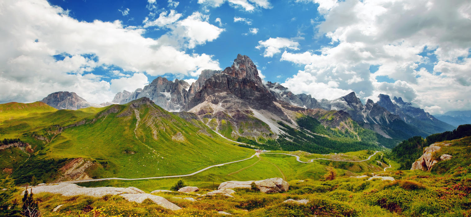 Una vista panoramica sulle cime frastagliate delle Dolomiti, con valli verdeggianti e una strada tortuosa sotto un cielo parzialmente nuvoloso, mostra perché questo sito del Patrimonio Mondiale dell'UNESCO è così impressionante.