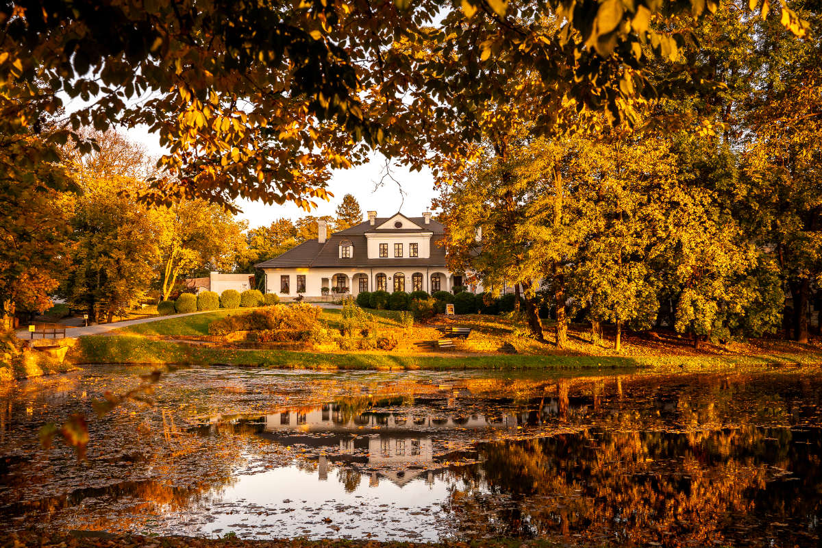 The large white house, reminiscent of Dwór Kombornia, is beautifully surrounded by autumn trees, reflecting in a pond under a clear sky.