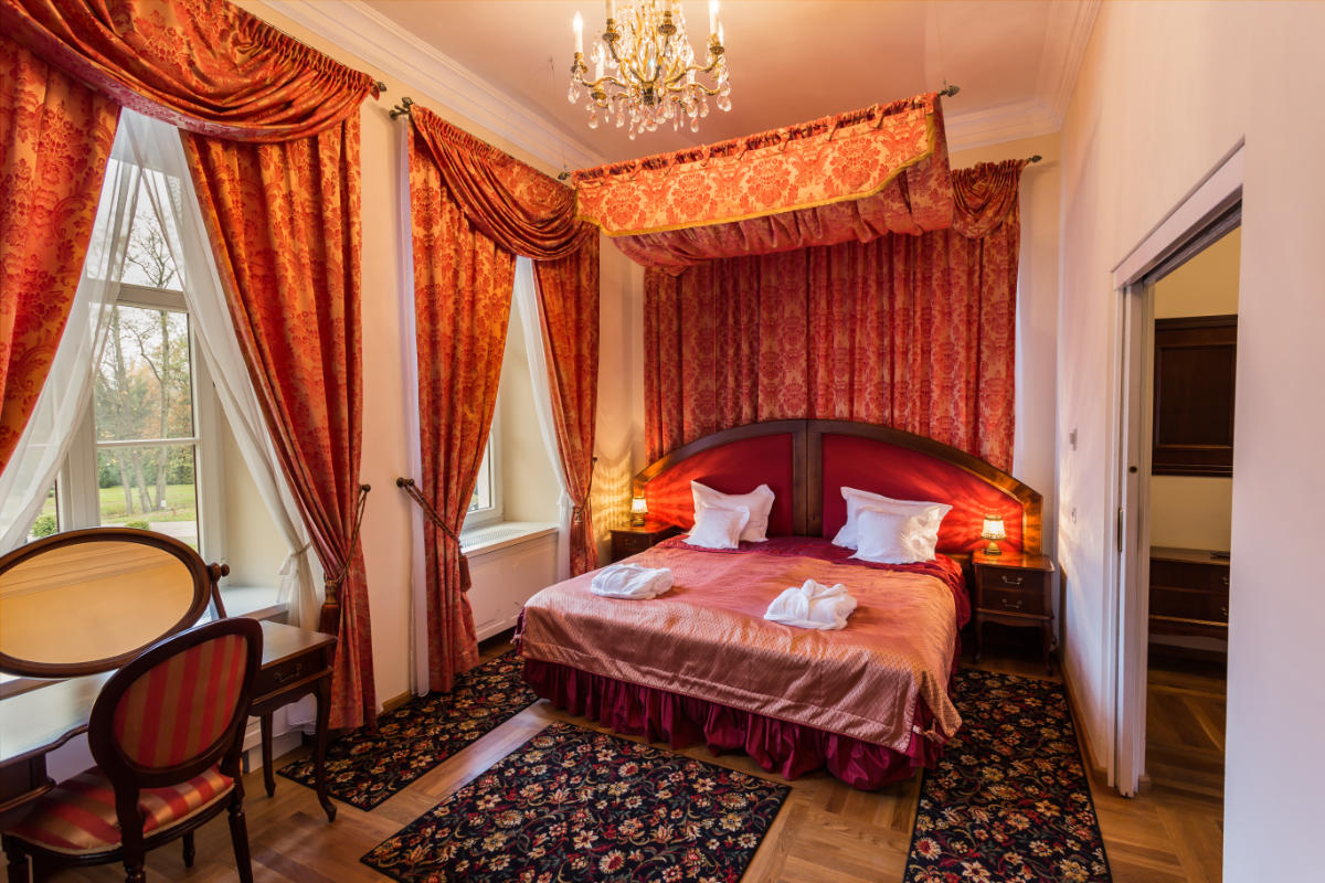 Elegant bedroom at Dwór Kombornia with a large bed, red and gold drapes, two windows, a chandelier, a desk with a chair, and patterned rugs.