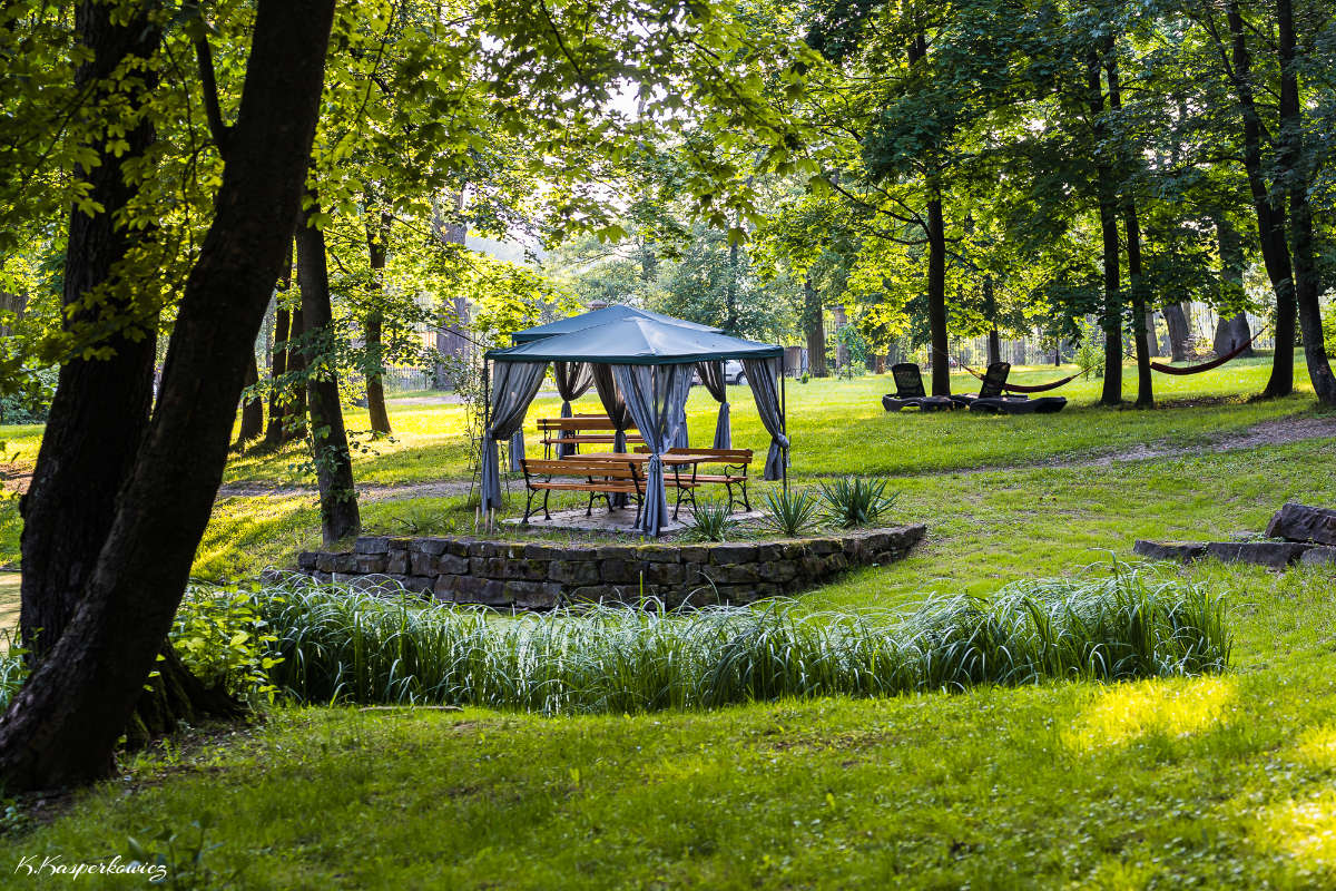 A gazebo with a green canopy and wooden picnic table sits in the lush grounds of Dwór Kombornia, surrounded by trees.