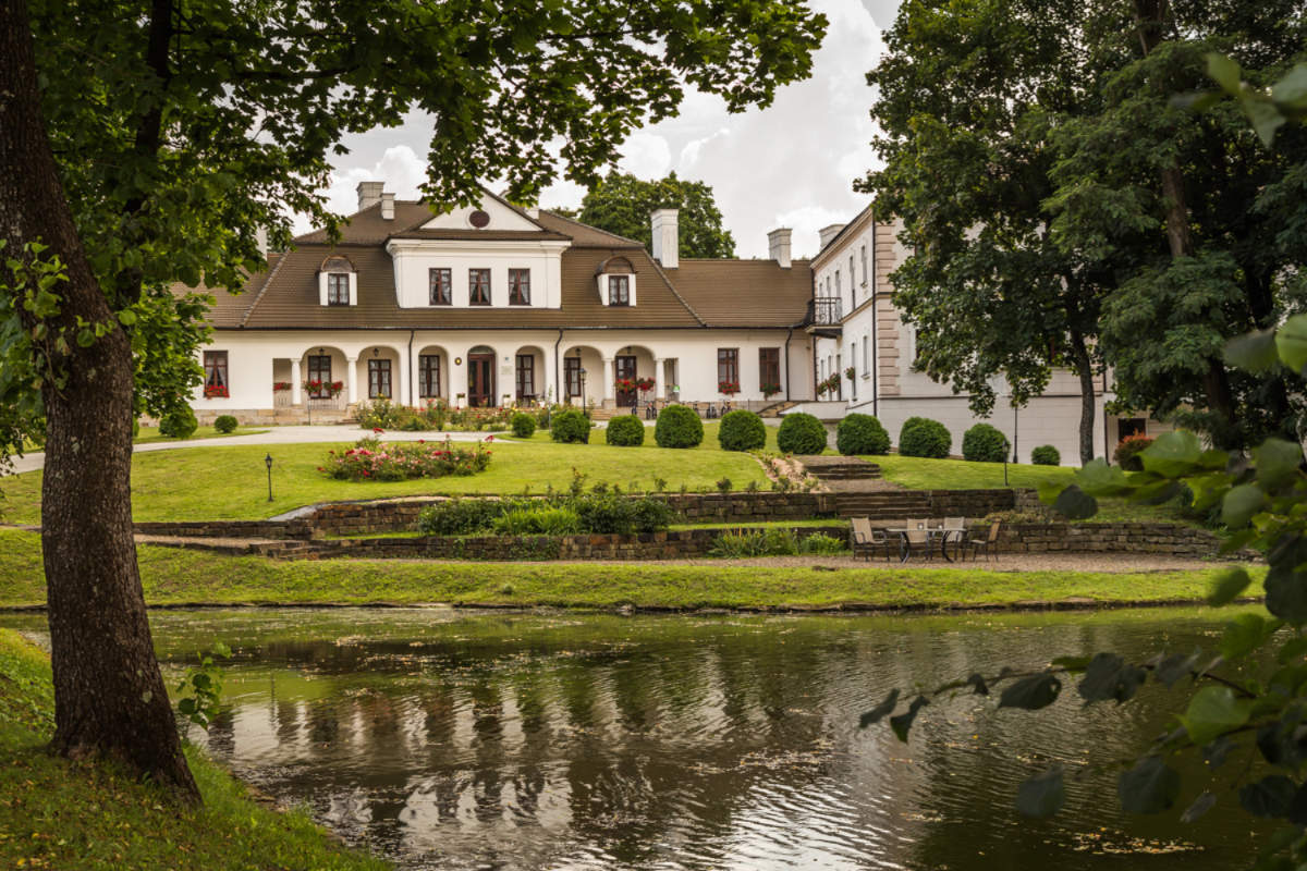 The grand estate of Dwór Kombornia, a splendid manor with a sloped roof and numerous windows, sits elegantly on a landscaped lawn beside a calm pond, surrounded by lush trees. This picturesque scene captures the essence of a dwór hotel in Poland.