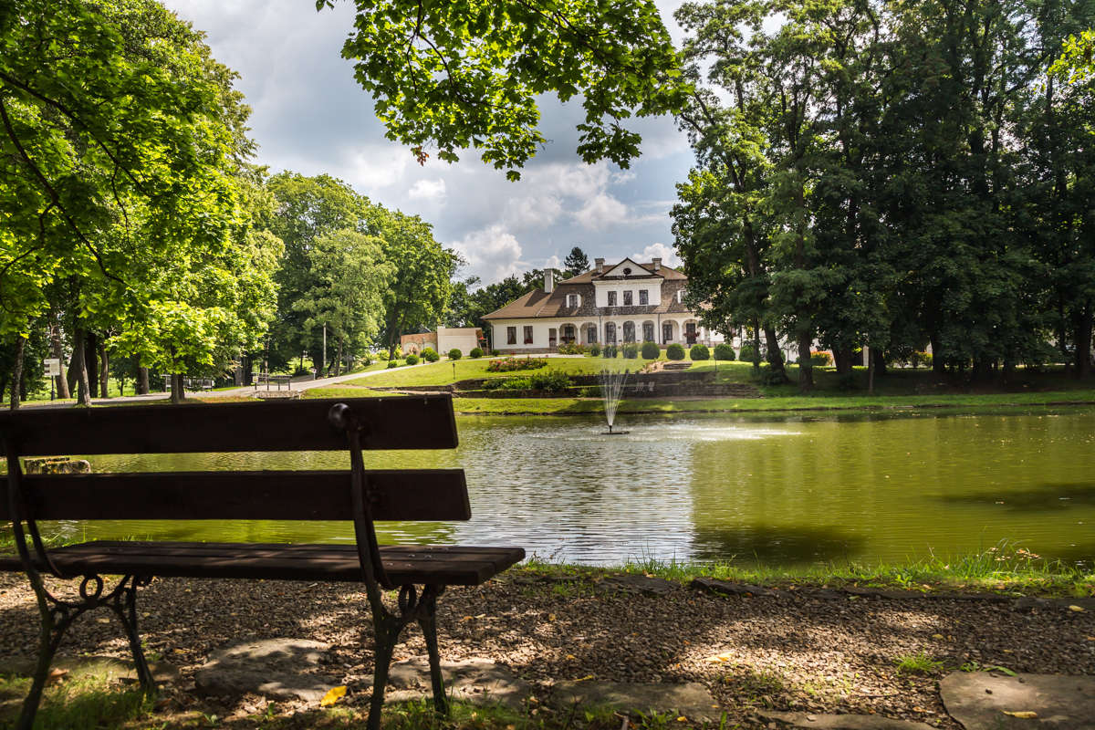 Scenic view of Dwór Kombornia, a large house by a pond surrounded by trees, with a bench in the foreground and a fountain in the water. This charming setting embodies the elegance of this historic dwór hotel in Poland.