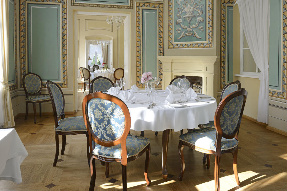 The elegant dining room at Dwór Kombornia features ornately decorated walls, a round table set for six, blue and gold upholstered chairs, and a stunning chandelier.