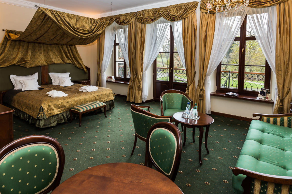 Experience an elegant room at Dwór Kombornia in Poland, featuring a large bed, green carpet, and classic wood furniture. Two tall windows with gold drapes illuminate a cozy seating area with a table and chairs, adding charm to this exquisite dwór hotel escape.