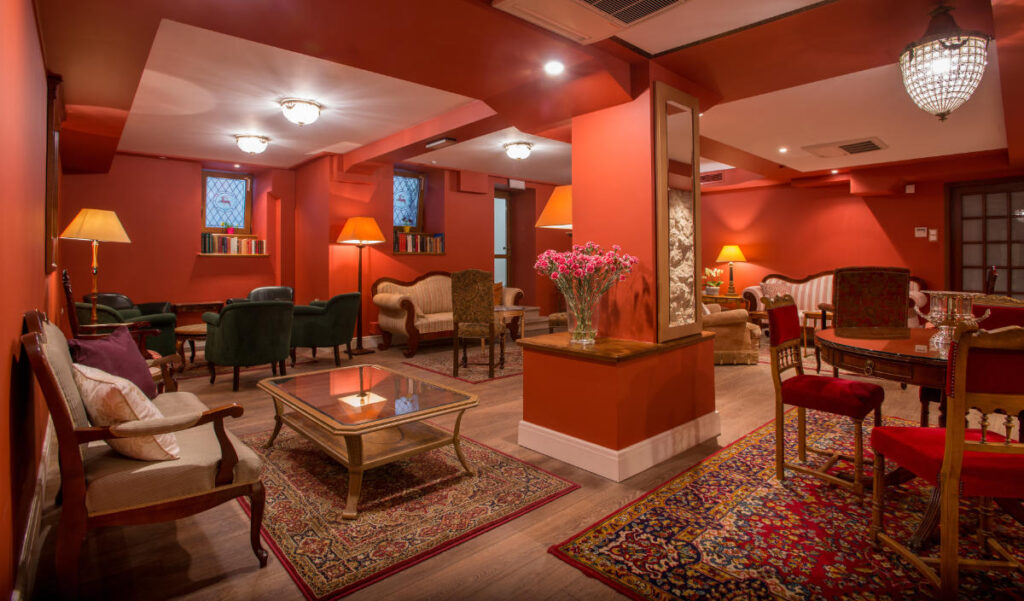 Il Club del libro degli alberghi storici: Fughe letterarie attraverso l'Europa 3 La lounge dell'Hotel Polski, illuminata in modo accogliente, presenta pareti rosse, diverse disposizioni di posti a sedere, tappeti persiani e un vaso di fiori decorativo su un tavolo.