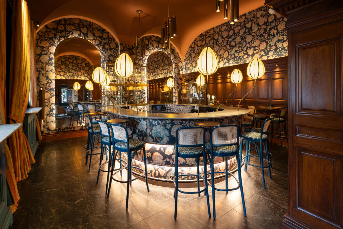 Elegante bar con bancone circolare e sgabelli alti, circondato da carta da parati floreale e illuminato da luci a sospensione a globo.