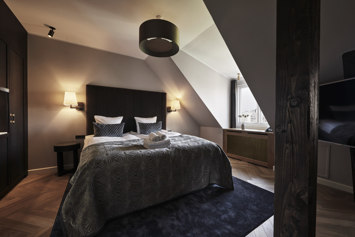 Una camera da letto elegante con letto matrimoniale, testata scura, biancheria da letto a fantasia, due lampade a parete, un grande tappeto, soffitto inclinato e una finestra che illumina la stanza.