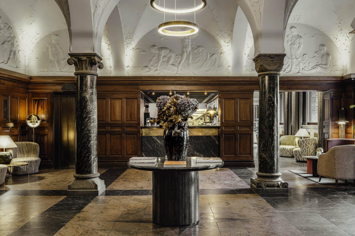 Die elegante Lobby des Hotel Ascot in Kopenhagen ist mit einem blumengeschmückten Marmortisch, einer verzierten Decke und Wandpaneelen aus Holz ausgestattet. Zwei Säulen und einladende Sitzecken vervollständigen diesen raffinierten Raum.