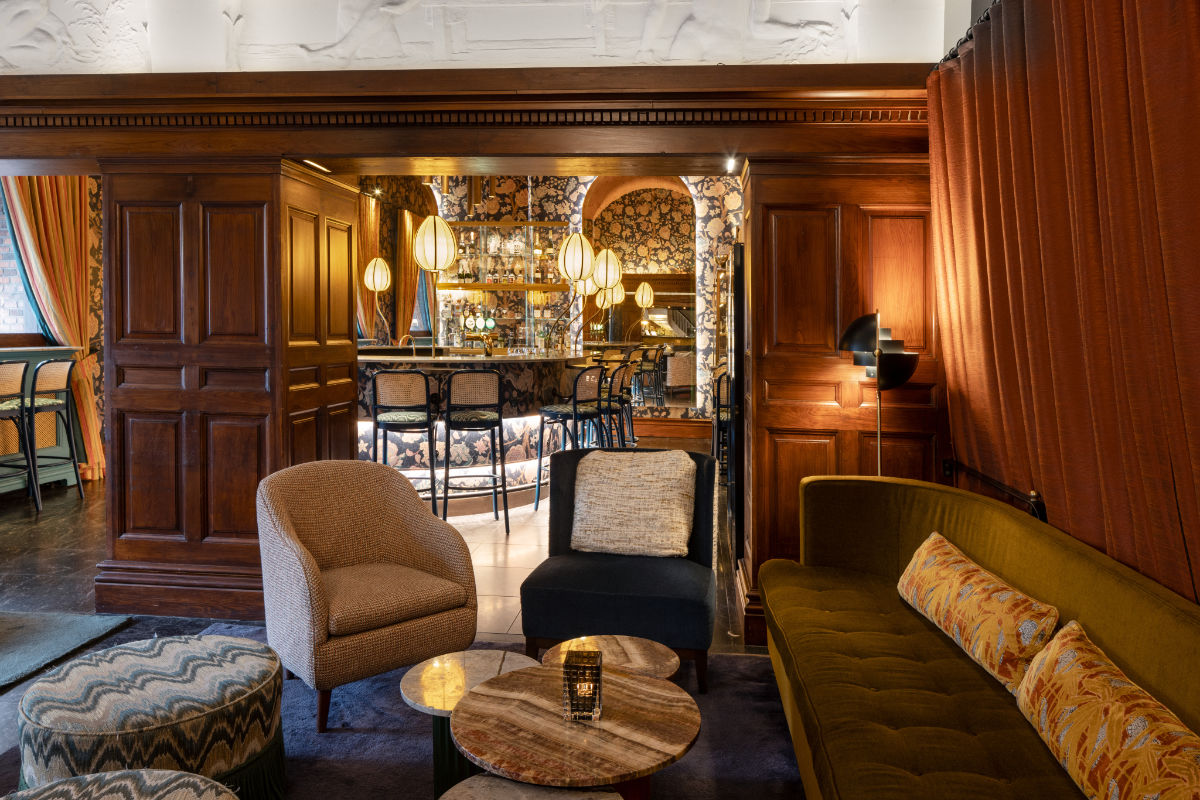 L'accogliente lounge dell'Hotel Ascot di Copenaghen è caratterizzata da sedute in velluto e carta da parati a motivi. Un bar con sgabelli e luci a globo aggiunge fascino allo sfondo, mentre l'illuminazione calda esalta i pannelli in legno e gli elementi decorativi, creando un'atmosfera invitante.