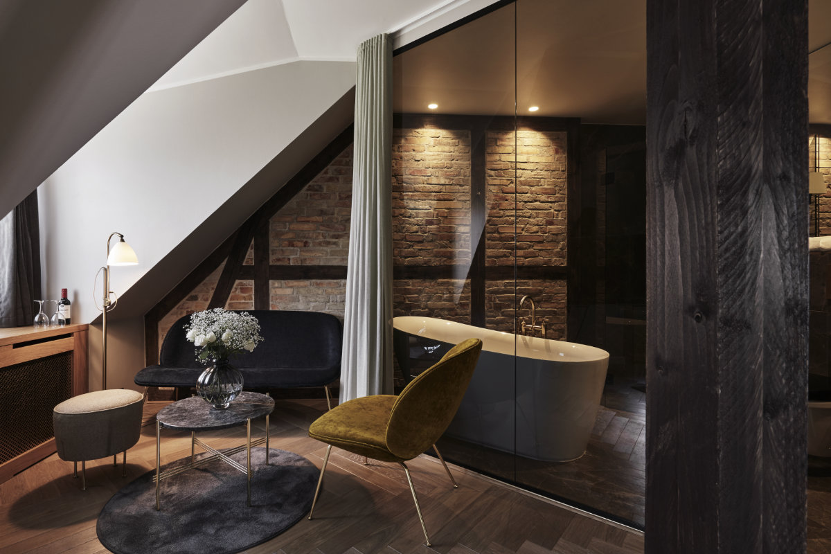 Moderno loft dell'Hotel Ascot, situato nel cuore di Copenaghen, con mattoni a vista e bagno con pareti in vetro e vasca. L'accogliente area salotto è dotata di divano, sedia e tavolini.