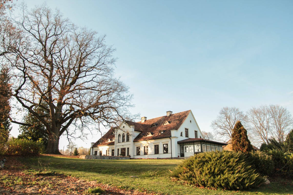The Mioduński Dworek is a large white house with a red roof, surrounded by lush trees and shrubs, all set against a clear blue sky.