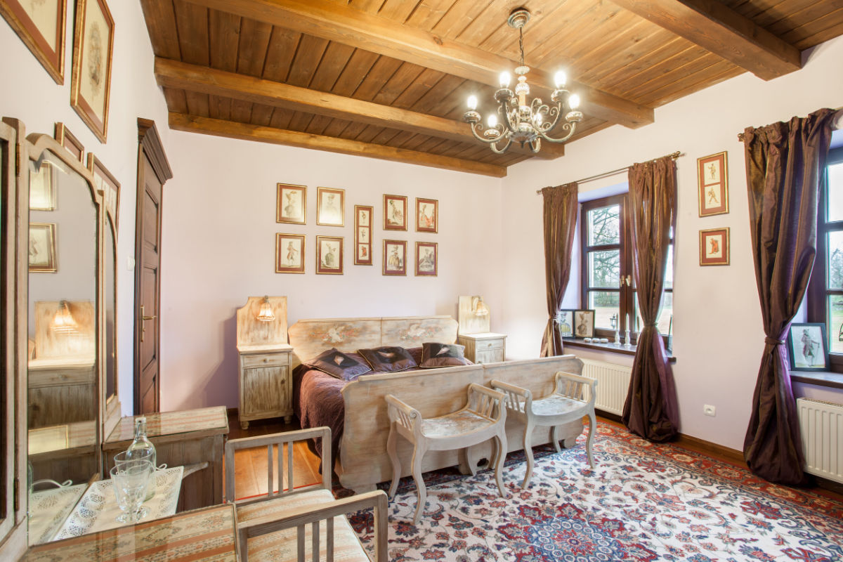 A rustic bedroom at Mioduński Dworek features a wooden ceiling, chandelier, vintage furniture, framed art, and patterned rugs. Two windows with curtains allow natural light to spill in, enhancing the room's charm.