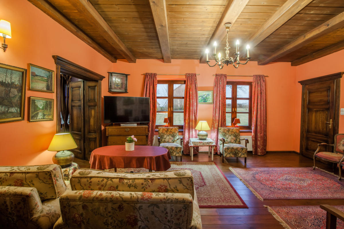 The cozy living room at Mioduński Dworek features warm orange walls and wooden ceiling beams. Floral sofas surround a round table, illuminated by elegant lamps and a chandelier. A TV, framed artwork, and red curtains on the windows complete the inviting setting.