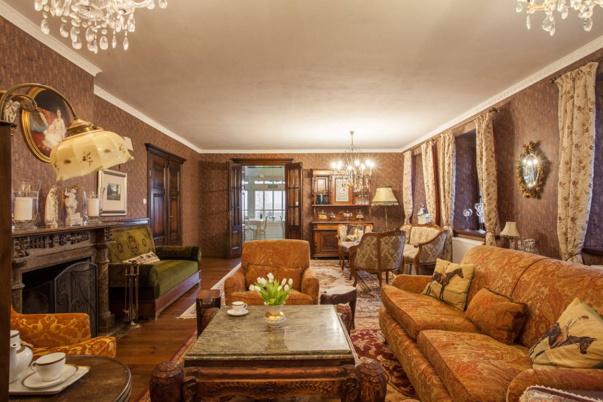 The Mioduński Dworek-inspired living room boasts vintage-style charm with patterned wallpaper, ornate chandeliers, and antique furniture. A marble-topped coffee table holds a vase of tulips, enhancing the room's elegant ambiance.