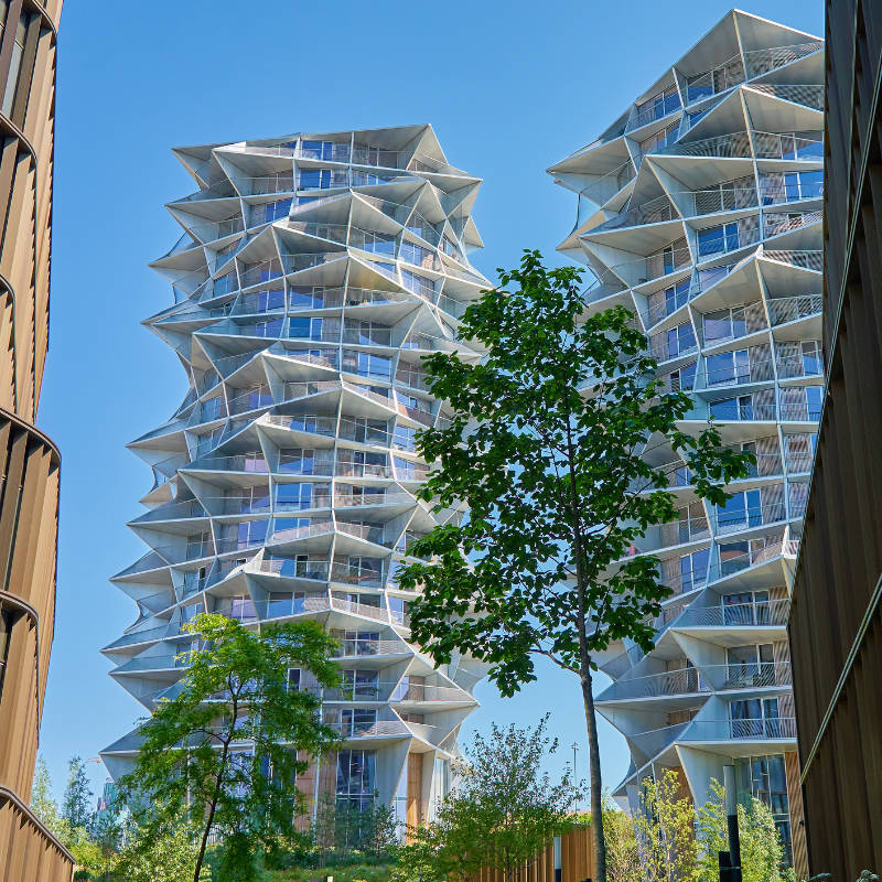 Dos modernos rascacielos de diseño geométrico con fachadas de cristal y metal, rodeados de árboles, bajo un cielo azul despejado.