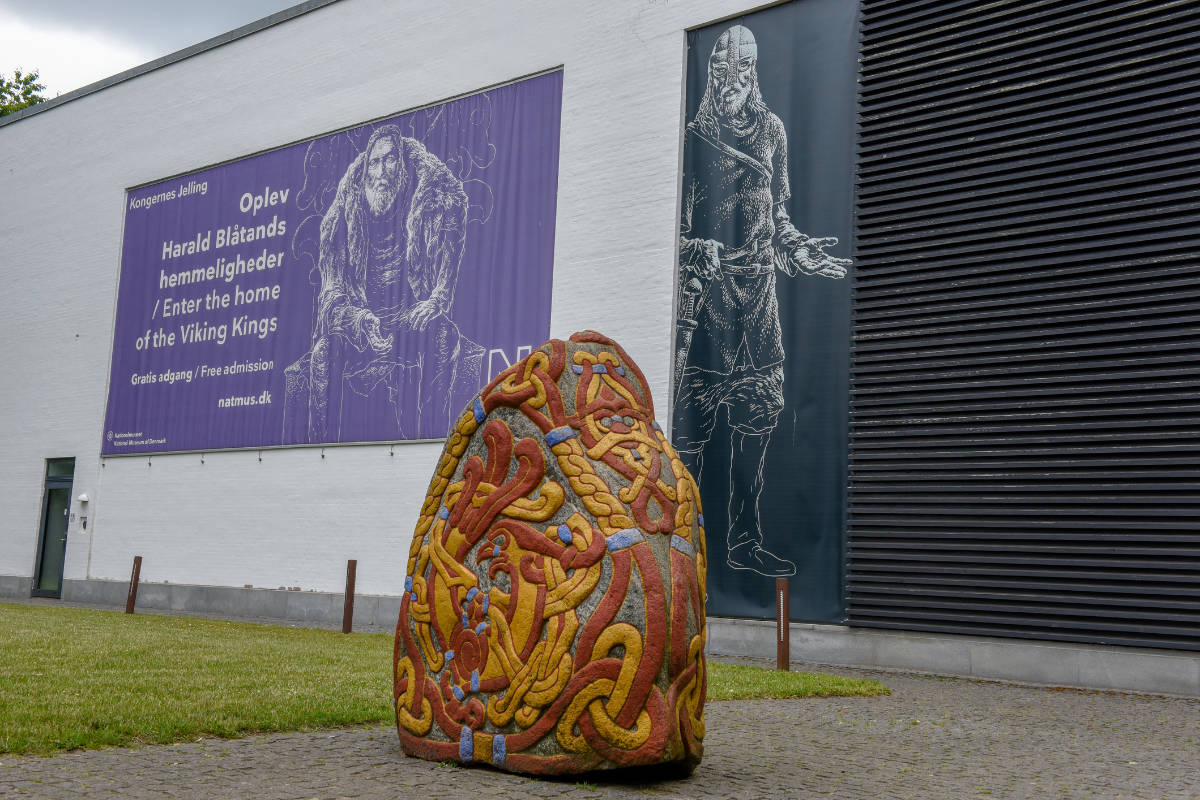 Colorida escultura de piedra rúnica en un camino empedrado frente a un edificio con un gran cartel mural de un personaje histórico.