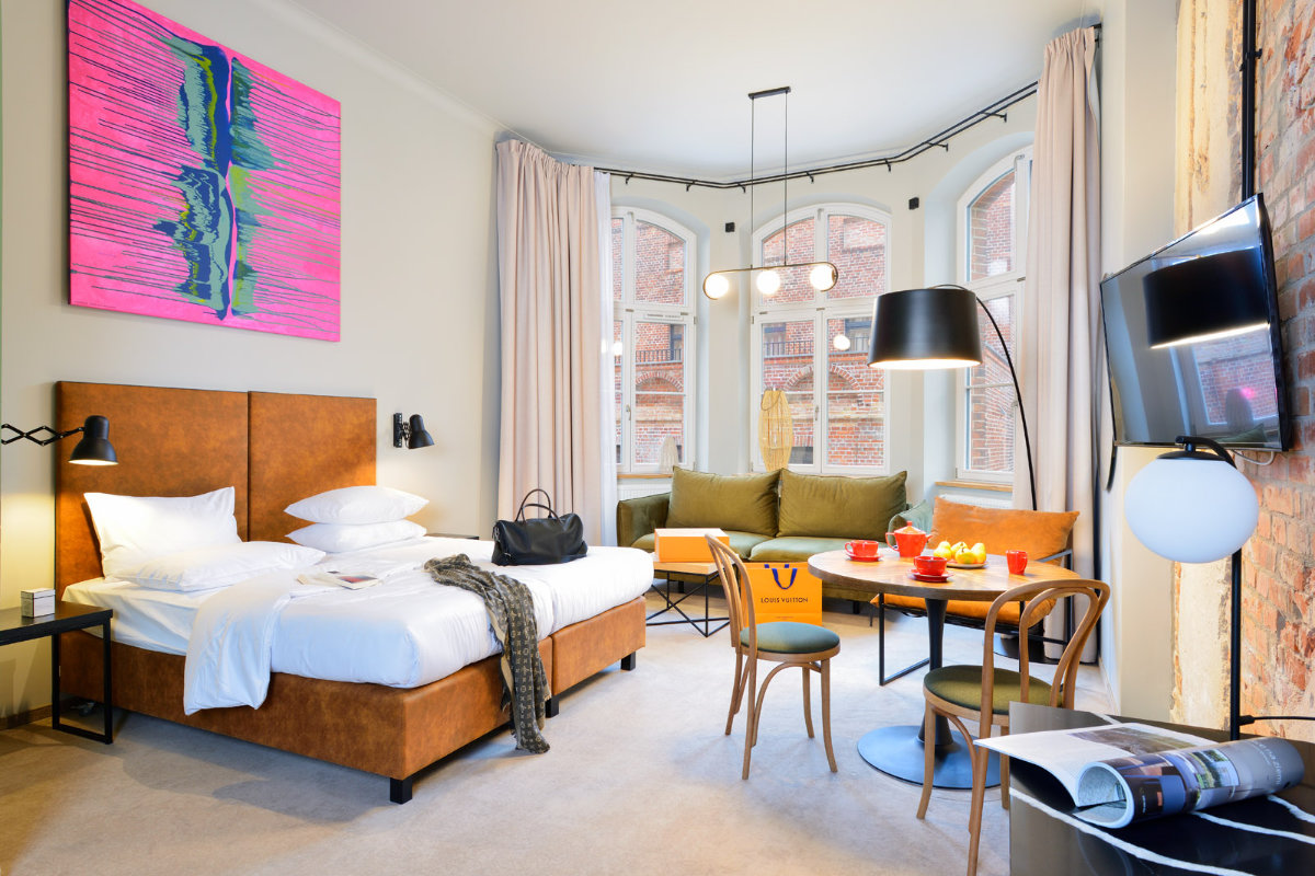Moderna camera d'albergo ispirata all'eleganza di Dwór Uphagena, dotata di letto matrimoniale, area salotto con grandi finestre, TV a parete e arte contemporanea. L'illuminazione luminosa esalta il mix di arredi in legno e pelle.