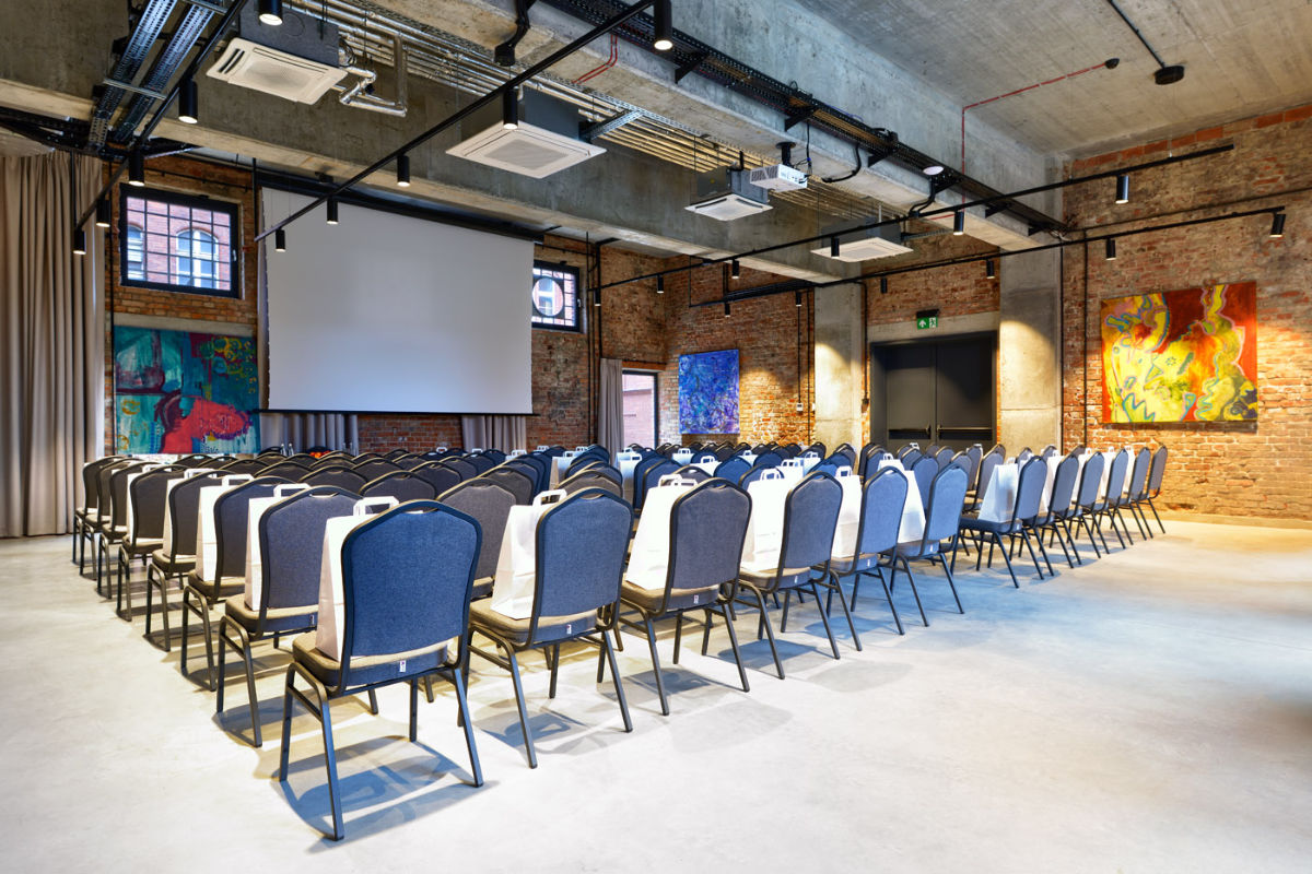 Una moderna sala conferenze con file di sedie di fronte a un grande schermo, che ricorda l'eleganza del Dwór Uphagena. Le pareti in mattoni a vista sono decorate con dipinti astratti colorati, creando un'atmosfera stimolante per gli incontri.