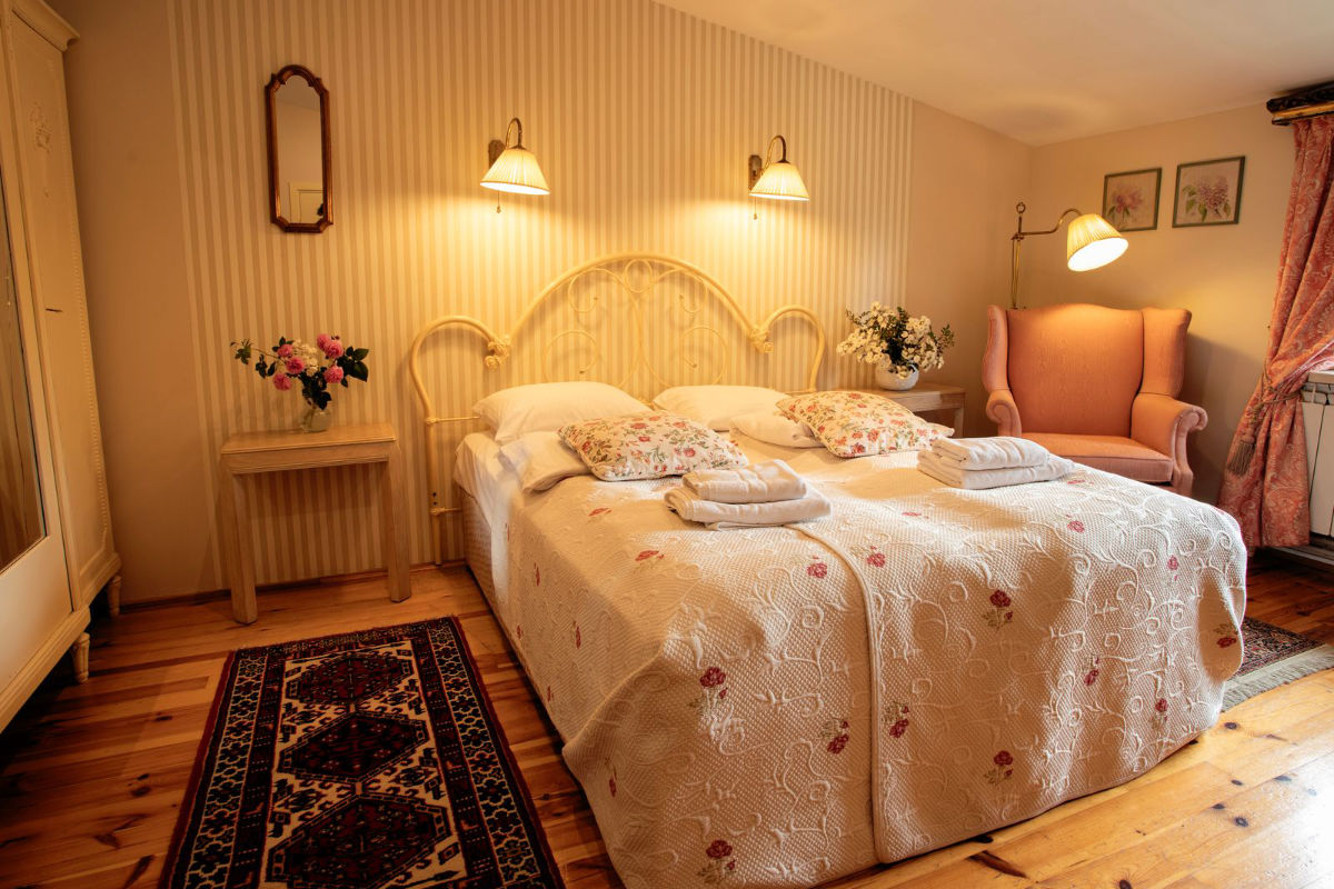 Un acogedor dormitorio con dos camas individuales juntas, una colcha de flores, cojines decorativos, mesillas de noche con flores, lámparas de pared, un gran sillón y una alfombra estampada sobre un suelo de madera.