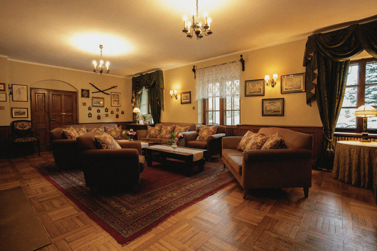 Un salón vintage con sofás marrones, una mesa de centro, cortinas adornadas, cuadros enmarcados y lámparas de araña en el techo sobre una alfombra estampada y suelo de madera.